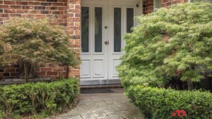 A brick house with a white front door, a small porch, and a well-maintained garden with various shrubs and a tree.