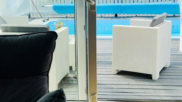 Une porte vitrée donne sur une terrasse extérieure avec une piscine, des meubles en osier blancs et, au fond, une clôture en bois peinte en blanc.