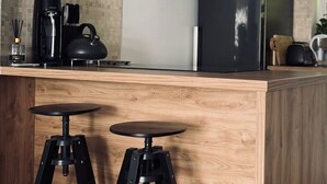 Une cuisine avec un plan de travail en bois, deux tabourets noirs, une machine à café et un réfrigérateur.