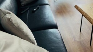 Un salon moderne avec un canapé en cuir noir, une table basse en bois et un fauteuil noir. Une télécommande est posée sur le canapé. La pièce est dotée d’une grande fenêtre donnant sur l’extérieur.