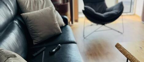Un salon moderne avec un canapé en cuir noir, une table basse en bois et un fauteuil noir. Une télécommande est posée sur le canapé. La pièce est dotée d’une grande fenêtre donnant sur l’extérieur.