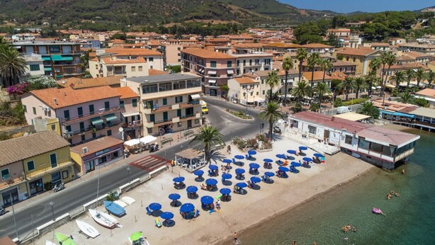 A coastal town with a beach area featuring umbrellas and boats, surrounded by residential buildings and mountains in the background. - Casa Francesca near center and beach (Porto Azzurro)