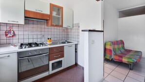A compact kitchen with white cabinets, a gas stove, and a built-in oven. There is a colorful chair in the adjacent room. - Casa Francesca near center and beach (Porto Azzurro)