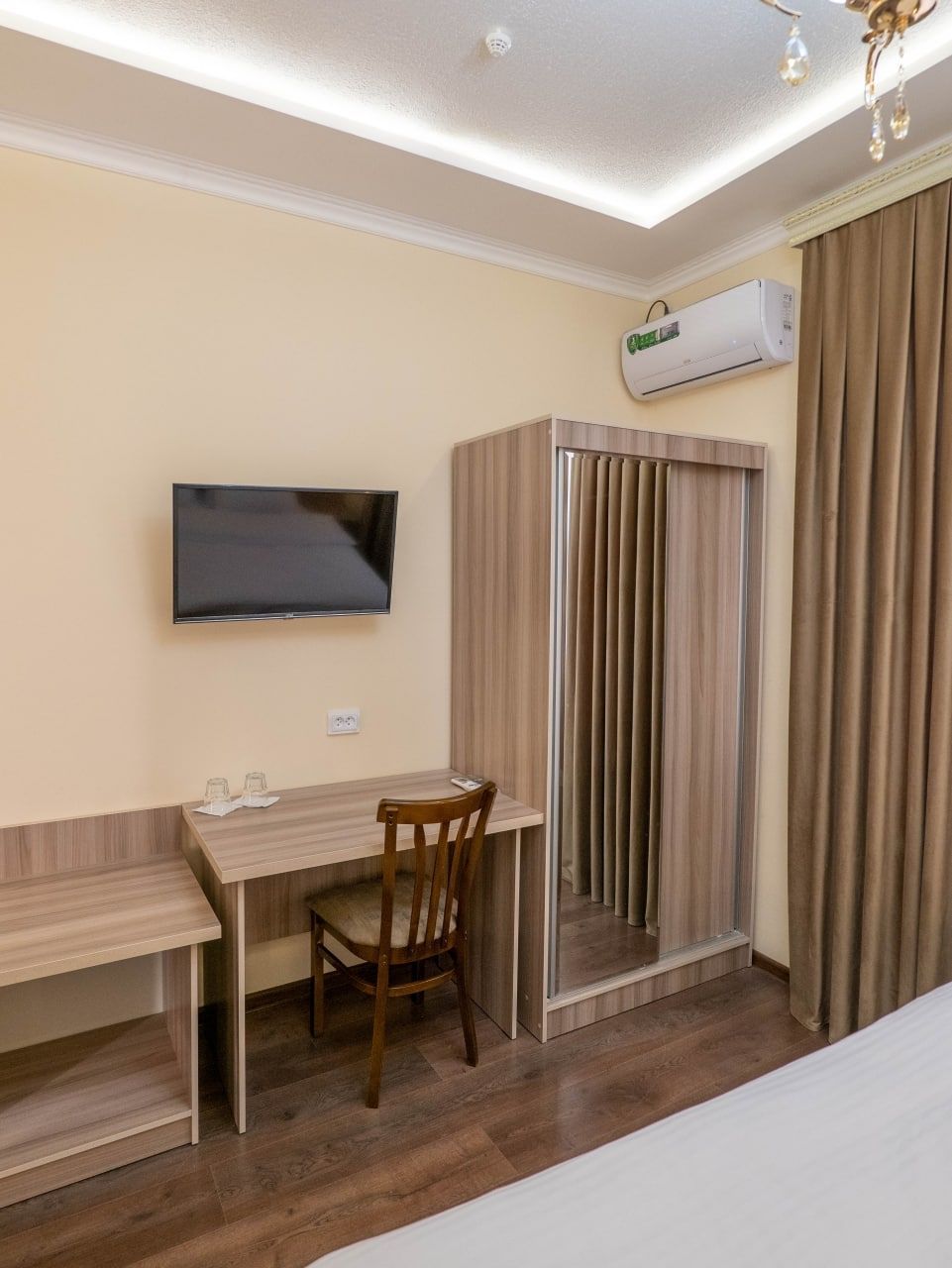 Una camera d'albergo con una scrivania in legno, una sedia, una televisione a parete, un condizionatore d'aria e una porta scorrevole con tende.