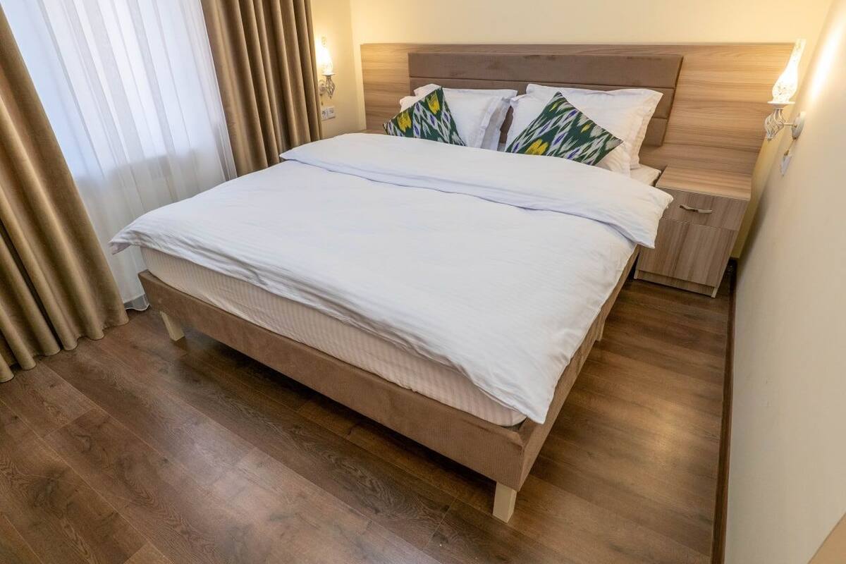 Un letto ben rifatto con una testiera in legno, un comodino con una lampada e una finestra con tende.