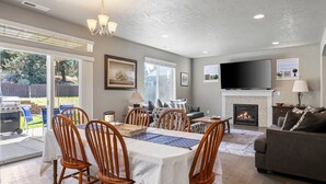 A spacious living room with a fireplace, a flat-screen TV, and a dining area with wooden chairs and a table. - Fantastic High Desert Retreat, Pet Friendly, Fenced Backyard w/ Covered Patio, Ping Pong, BBQ (Bend)
