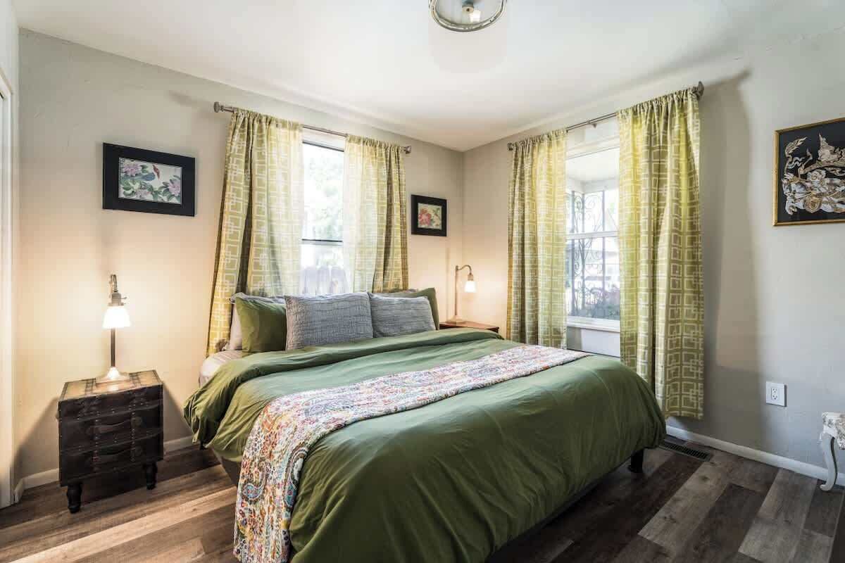 Et soveværelse med seng, gardiner, et natbord, en lampe og et billede på væggen.