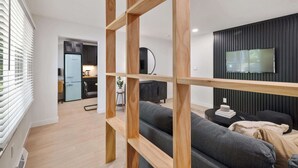 Un salon moderne avec un canapé gris, des étagères en bois, un téléviseur fixé sur un mur foncé, et une cuisine en arrière-plan.