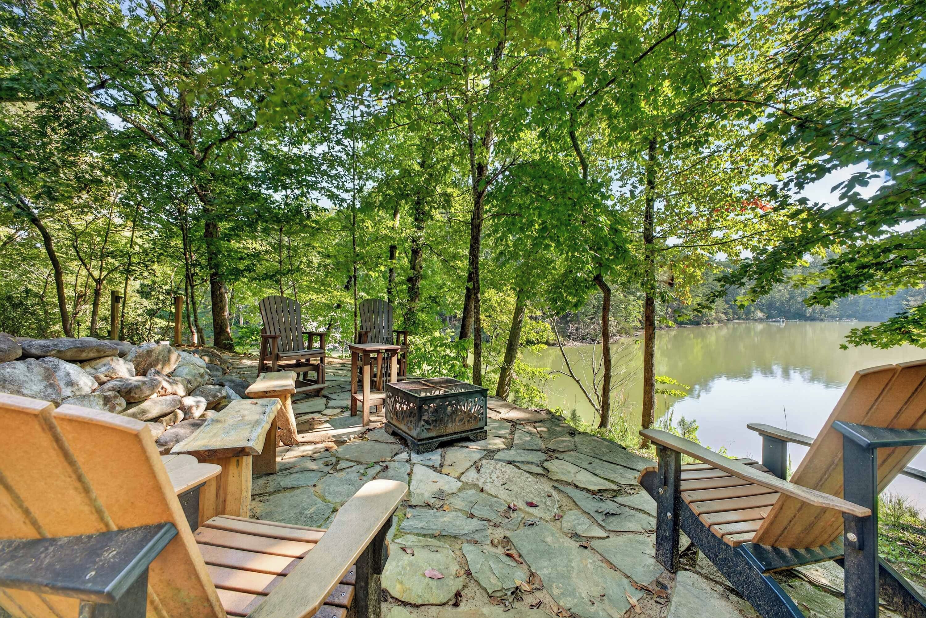 An outdoor seating area with wooden chairs and tables, surrounded by trees and overlooking a lake.