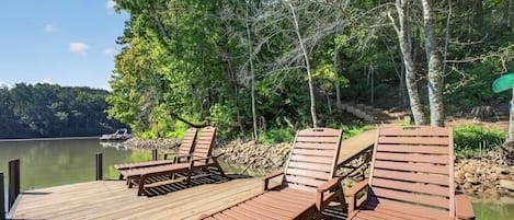 A wooden deck with lounge chairs and a dock extending into a calm lake, surrounded by trees.