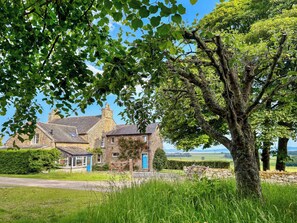 A stone house with a blue door, surrounded by lush greenery and a large tree. - The Annex at Mains House (Tain)