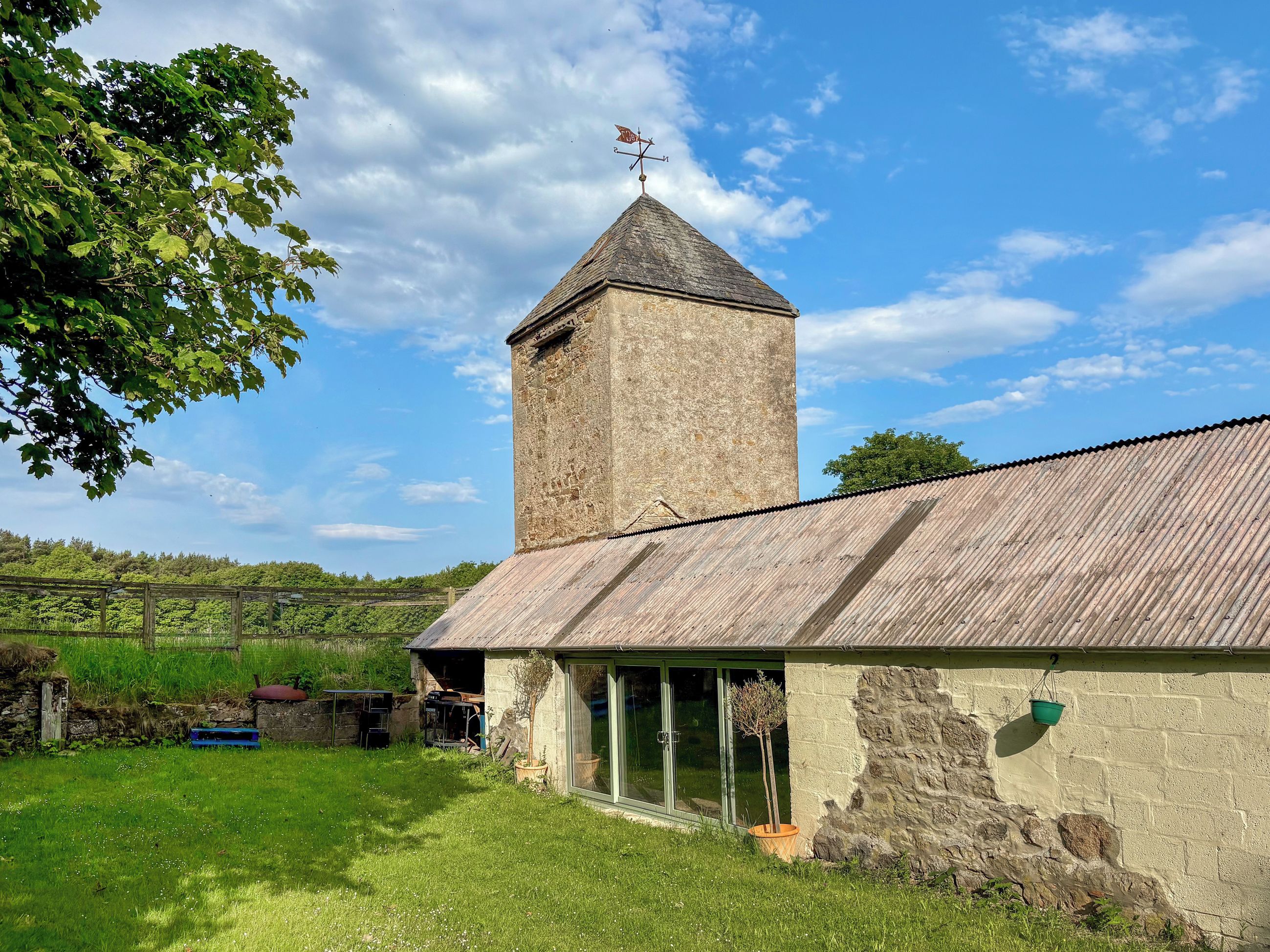 A stone tower with a weather vane, a wooden building with a corrugated metal roof, and a grassy yard with a fence and potted plants.