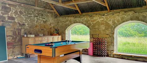 Une table de billard dans une pièce aux murs en pierre et aux poutres apparentes, de grandes fenêtres donnant sur un paysage verdoyant, et un meuble en bois.