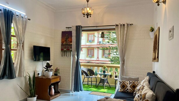 A living room with a sofa, a TV, a ceiling fan, and a balcony with a view of buildings. - 1bhk flat with pool view 5 mins walk to Candolim beach in a prime location. (Calangute)