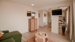 A modern living room with a green sofa, a wooden cabinet, a round table with remotes, and a TV mounted on the wall.