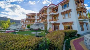 Un hotel de varios pisos con balcones, un jardín cuidado y montañas al fondo.