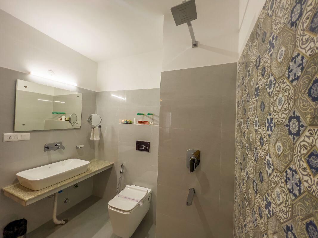 A bathroom with a white sink, mirror, and toilet. There is a shower area with a patterned wall and a door.