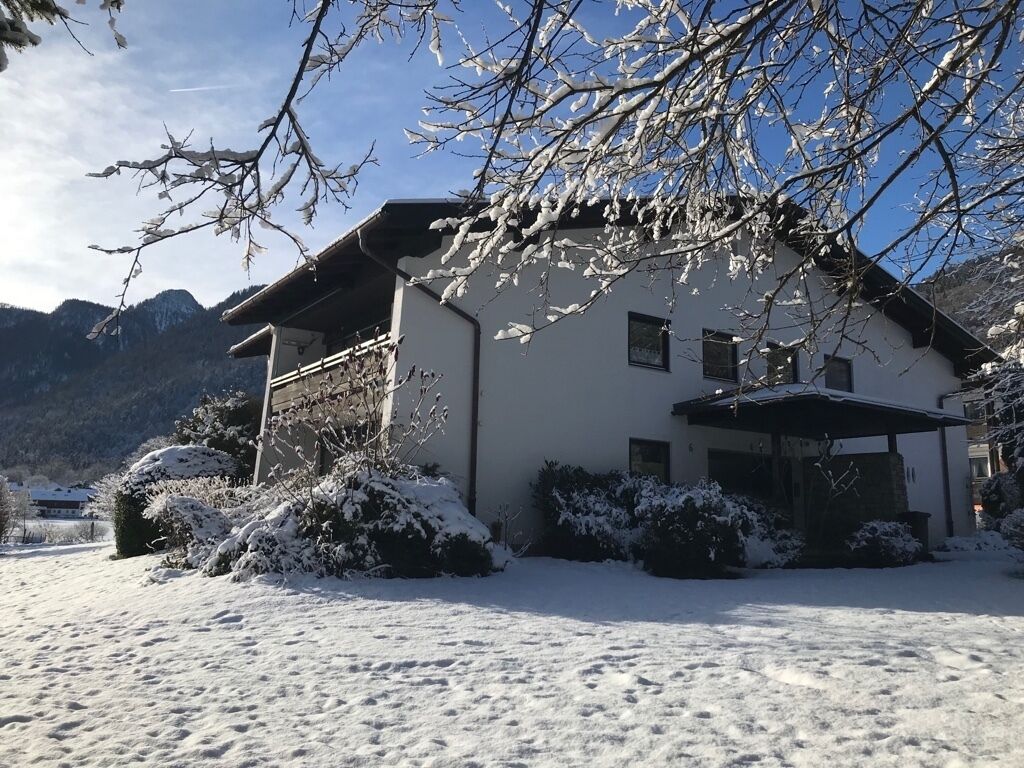 Una casa con techo inclinado, rodeada de arbustos cubiertos de nieve y un paisaje montañoso de fondo.