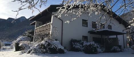 Una casa con techo inclinado, rodeada de arbustos cubiertos de nieve y un paisaje montañoso de fondo.