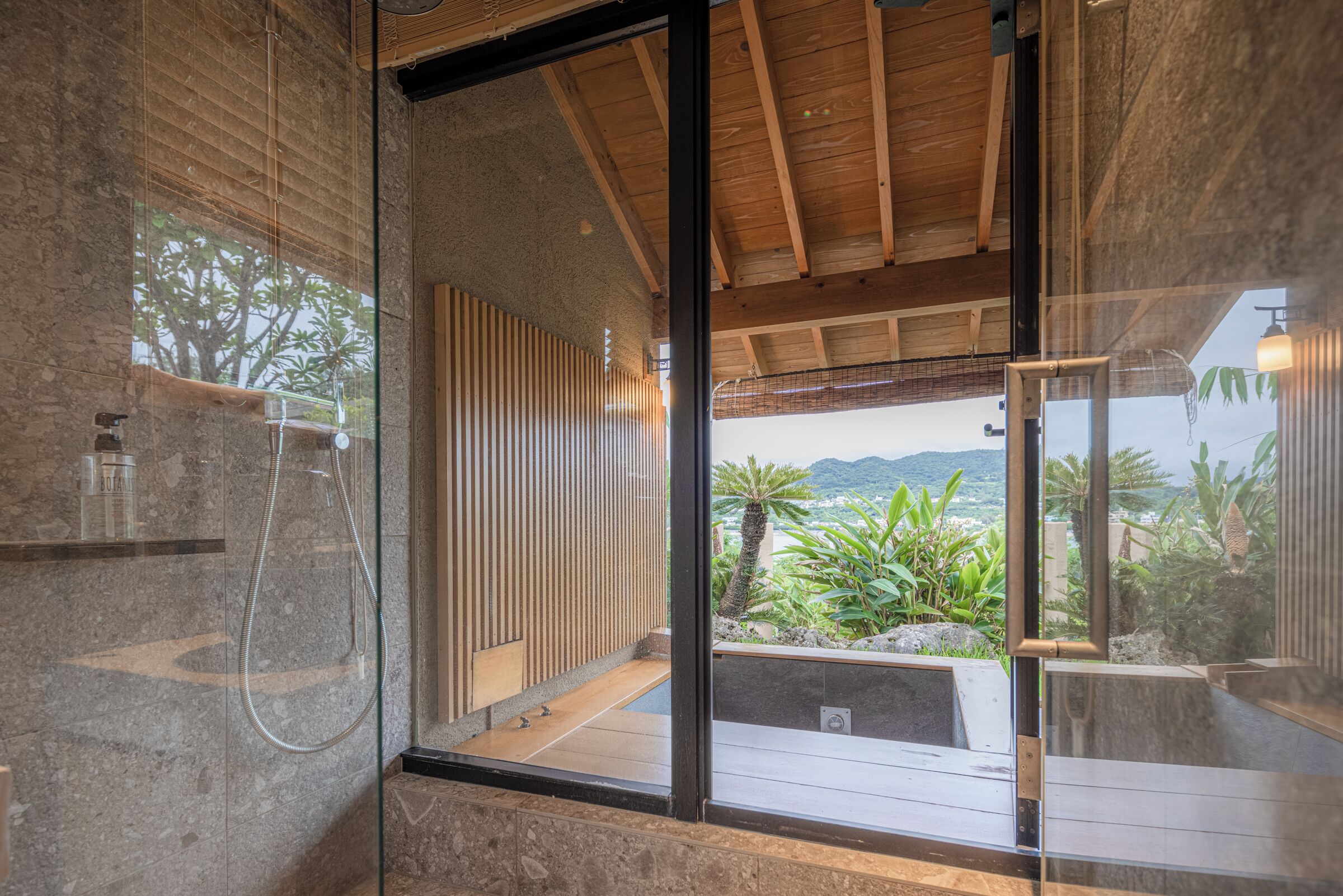 A modern bathroom with a glass shower enclosure, wooden ceiling beams, and a view of a tropical landscape.