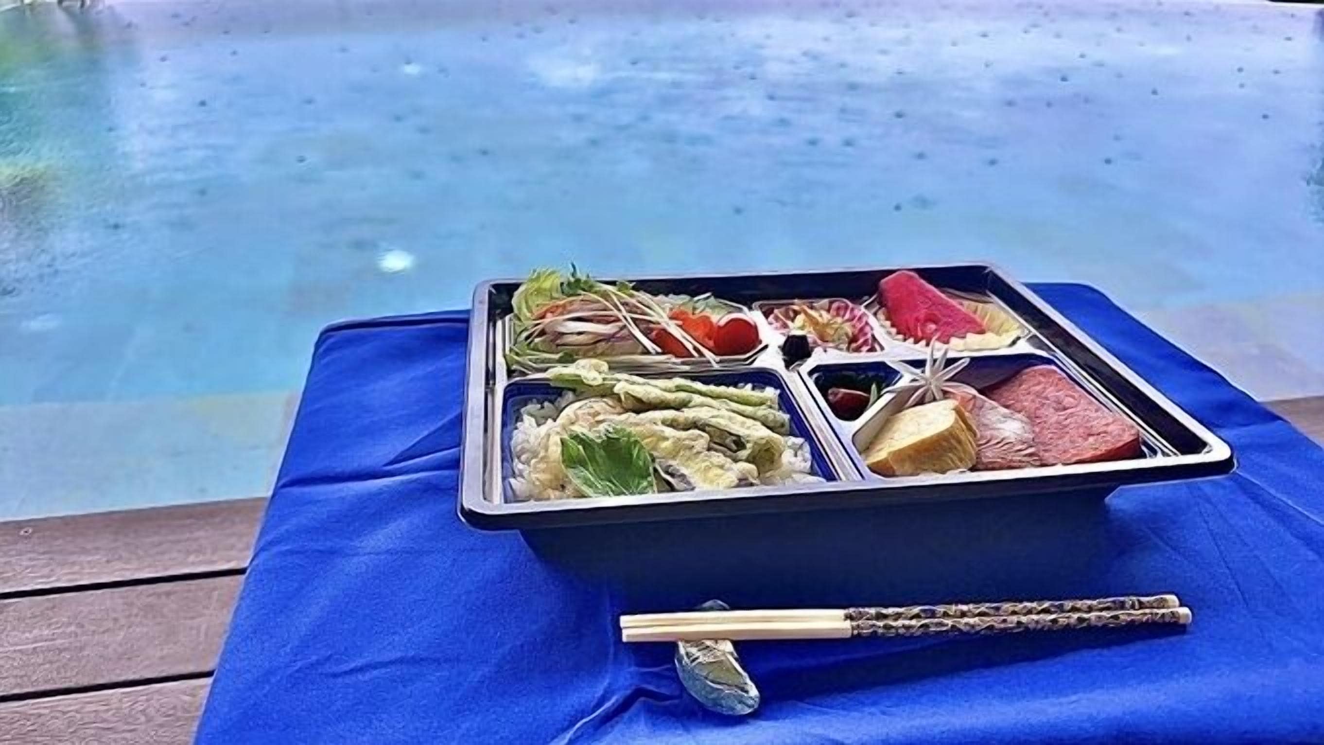 Una caixa de bento amb diversos aliments sobre un drap blau a la vora d'una piscina.