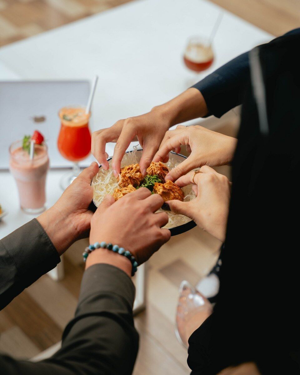 A group of people sharing a meal around a table with a laptop and drinks in the background.