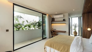 A modern bedroom with a large bed, a desk, and a view of a balcony with plants. - Makia Penthouse by Alfred in Bali (Canggu)