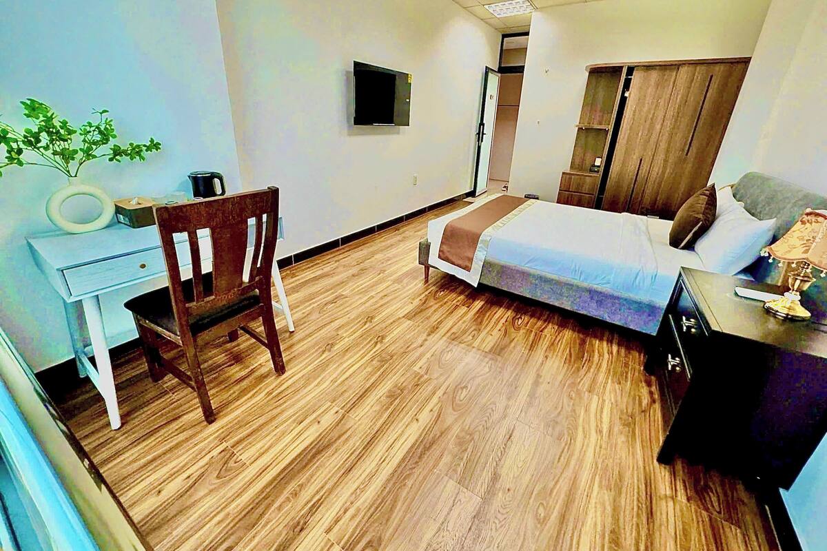 Een hotelkamer met een houten vloer, een bed met een bruin en wit dekbed, een bureau met een stoel, een televisie aan de muur en een kledingkast.