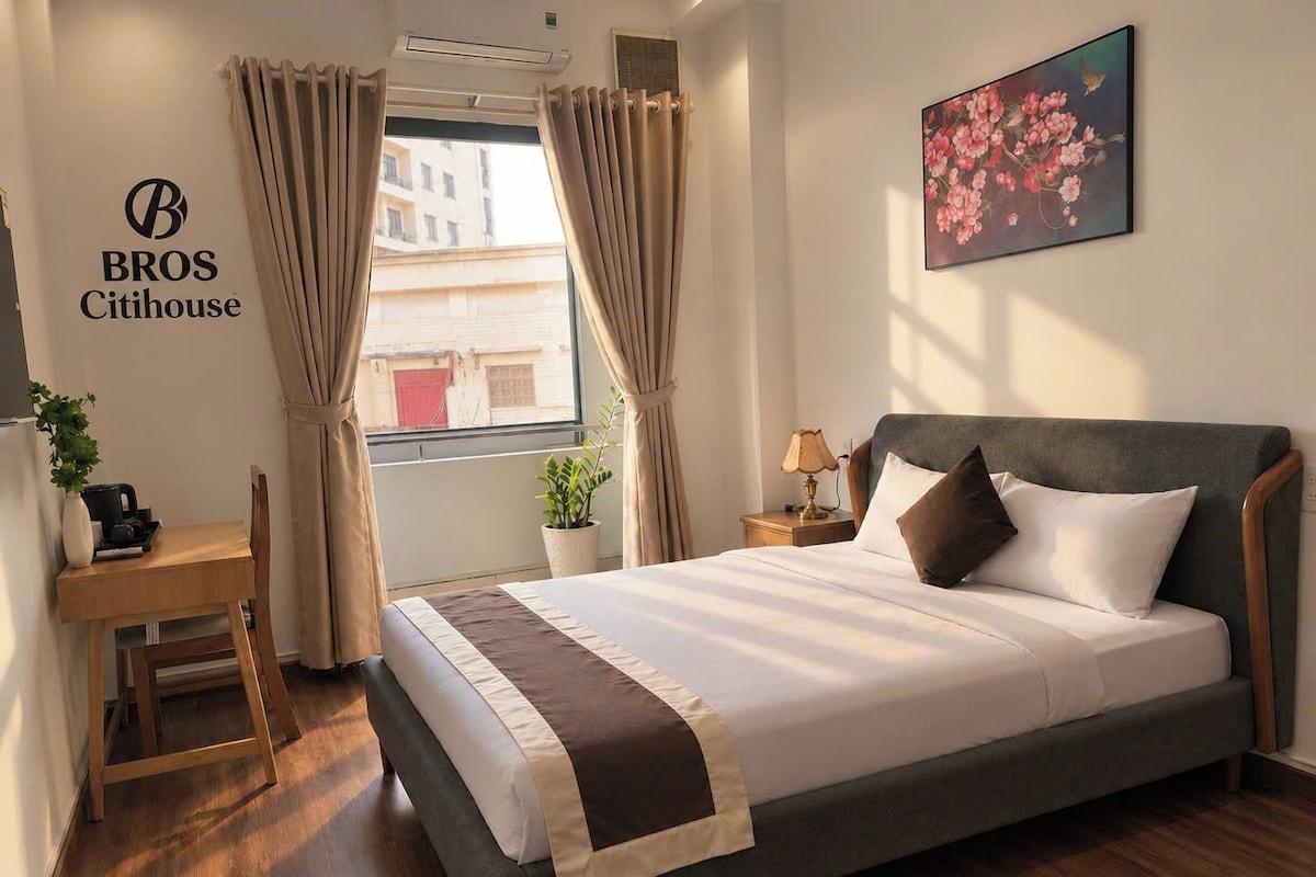 Una habitación de hotel con cama, mesita de noche, lámpara y ventana con cortinas.