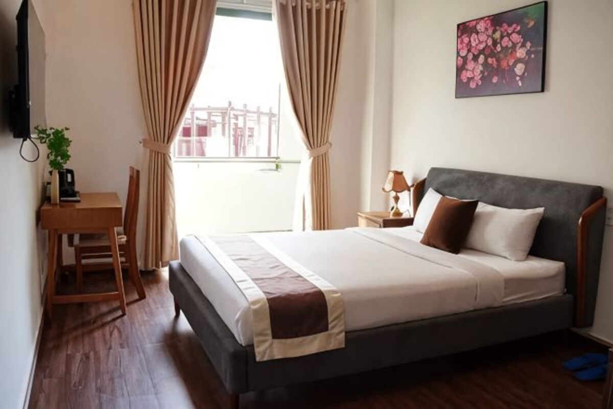 A bedroom with a bed, a desk, a chair, a nightstand, a lamp, and a window with curtains.
