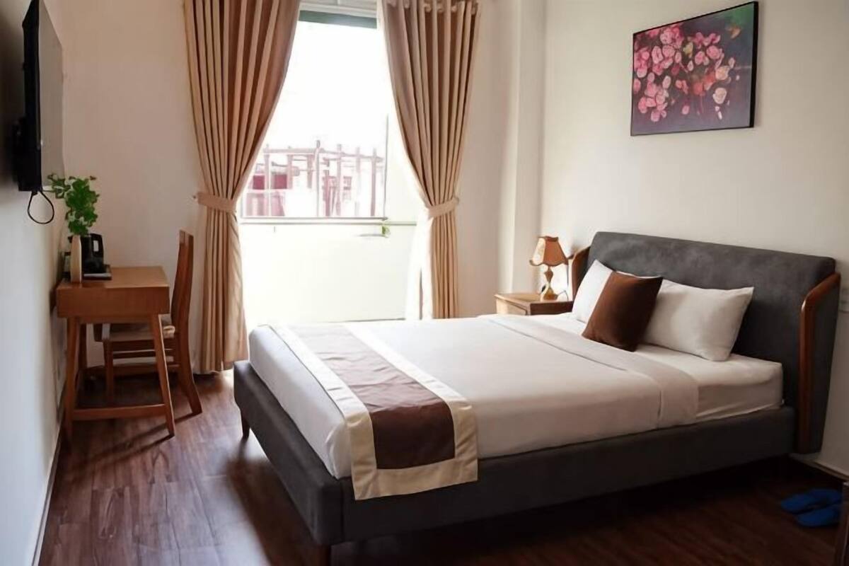 Een slaapkamer met een bed, een bureau, een stoel, een nachtkastje, een lamp en een raam met gordijnen.