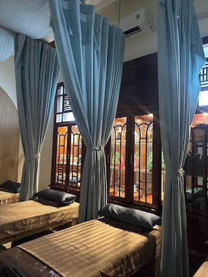 Habitación con dos camas, cada una con almohada y manta. Cortinas atadas en la parte superior y ventana con vista a un espacio exterior.