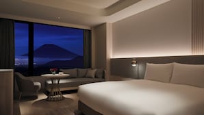 Habitación de hotel moderna con una cama grande, un área de descanso con una mesa y vistas a una montaña al atardecer.