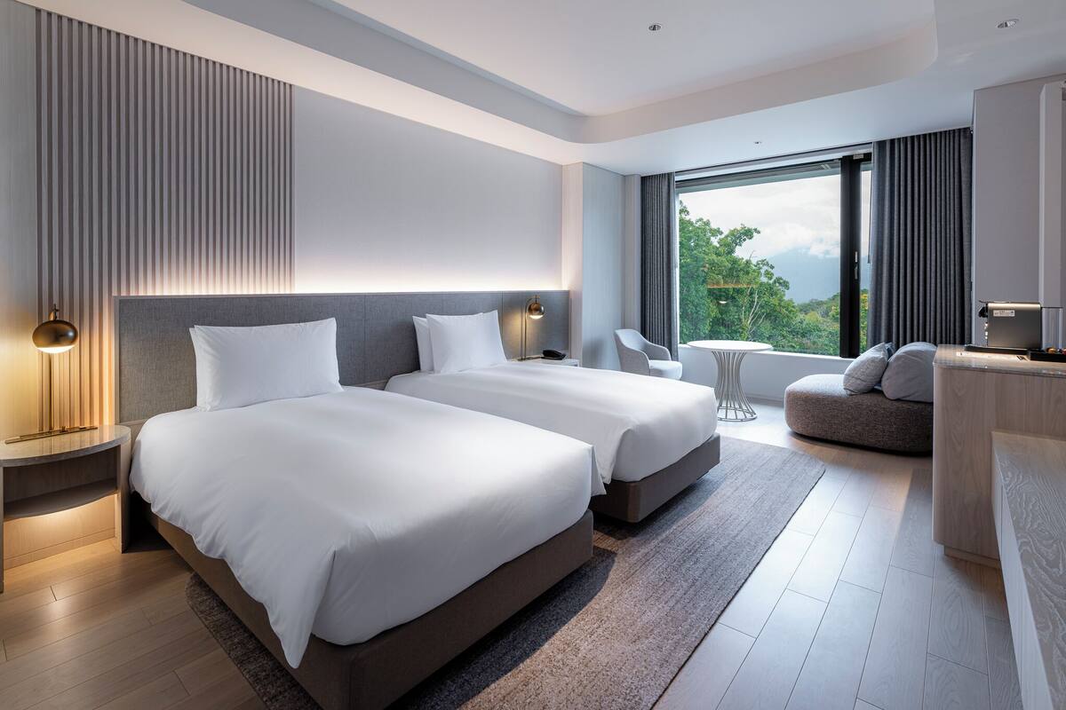 A modern hotel room with two beds, a small seating area, and a large window offering a view of the outdoors.