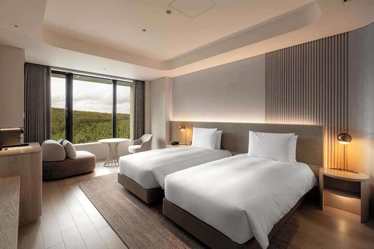 A modern hotel room with two beds, a desk, a sofa, and large windows offering a view of the outdoors.