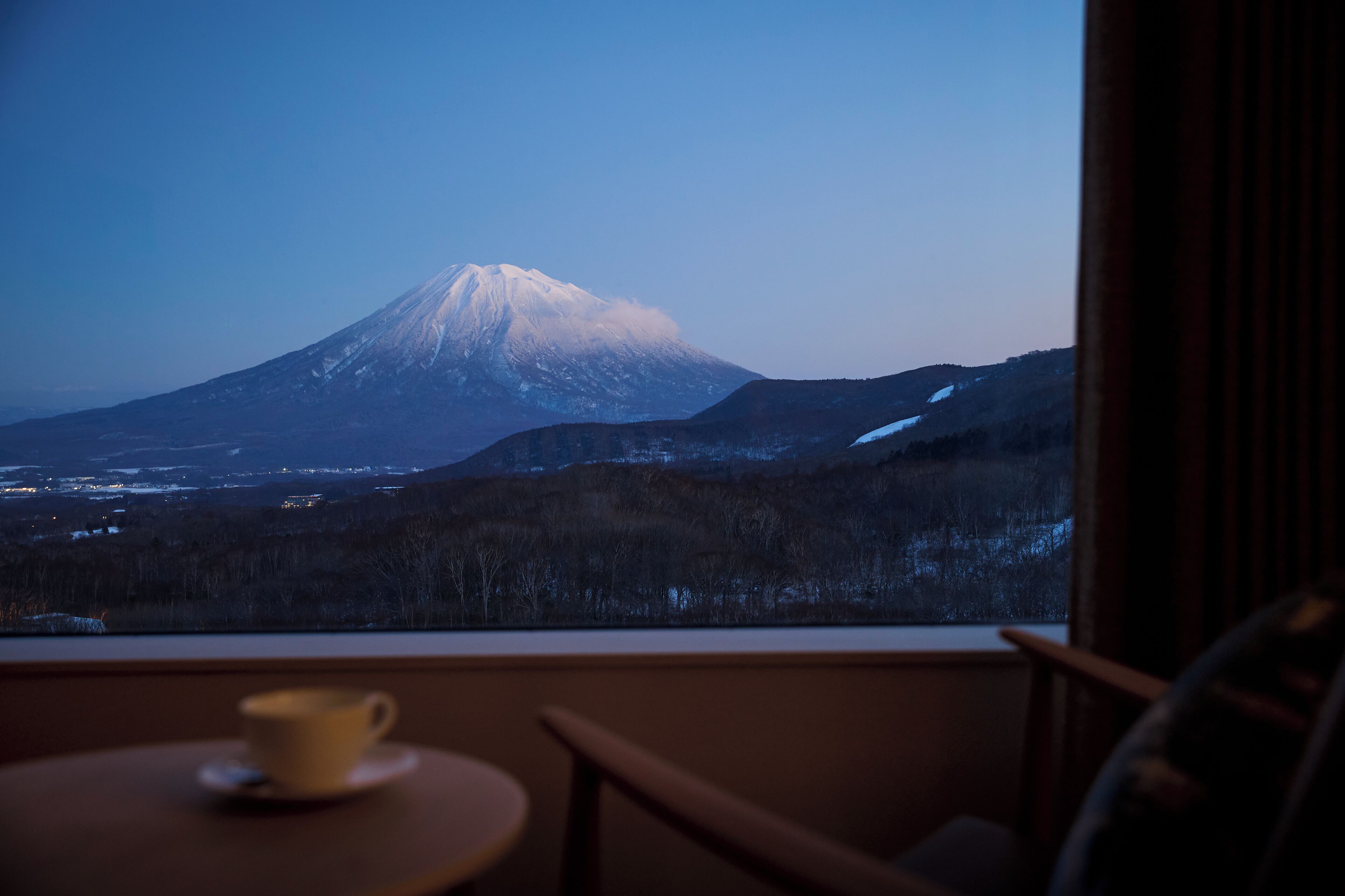 A view of a snow-capped mountain from a window, a cup on a table, and a chair.