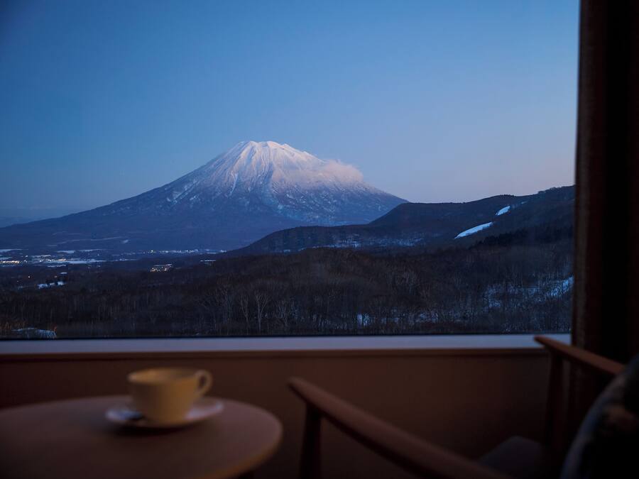 A view of a snow-capped mountain from a window, a cup on a table, and a chair.