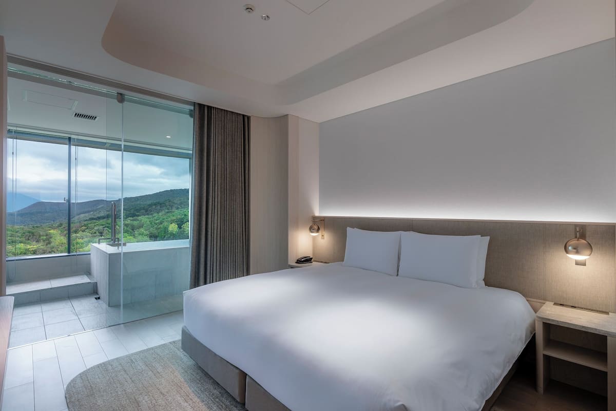 A modern hotel room with a large bed, a glass wall with a view of the mountains, a small seating area, and a nightstand with a lamp.