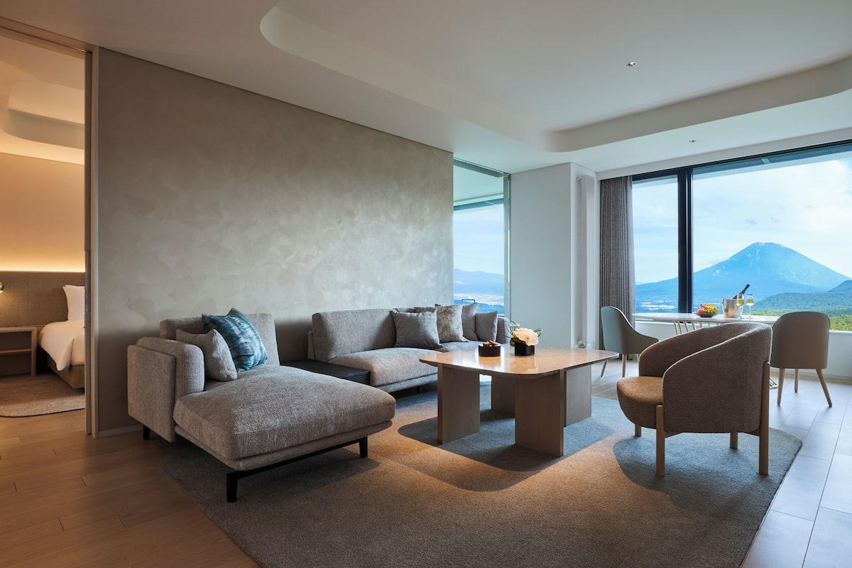 A modern living room with a sofa, armchair, and coffee table, large windows with a mountain view, and a dining area with chairs.
