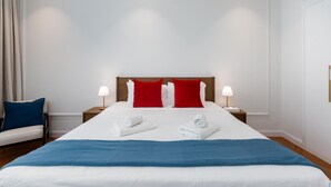 Een hotelkamer met een groot bed, twee rode kussens, wit beddengoed, twee bedlampjes en een blauwe plaid.