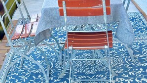 En terrasse med et bord dækket til to, omgivet af metalstole, en gulvtæppe med et mønster og en glasbeholder med en plante.