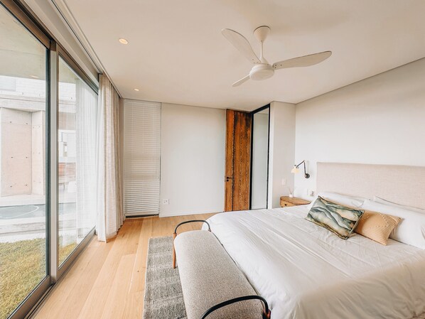 A modern bedroom with a large bed, a ceiling fan, a wooden door, and a view through a large window.