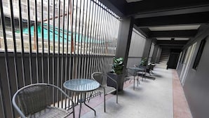 Un pasillo con sillas y mesas de metal, una planta en maceta y vista a una obra en construcción a través de barrotes.