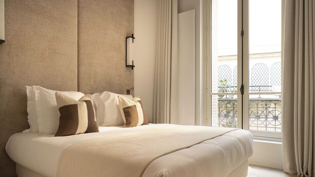 Ein ordentlich bezogenes Bett mit einer beigen Bettdecke und Kissen, einer an der Wand befestigten Lampe und Blick auf einen Balkon mit Metallgeländer.