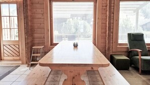A wooden cabin interior with a large dining table, two windows with blinds, a green chair, and a small stool. - Villa Persikka, Himoslaakso Ski-in/out (Keski-Suomi)