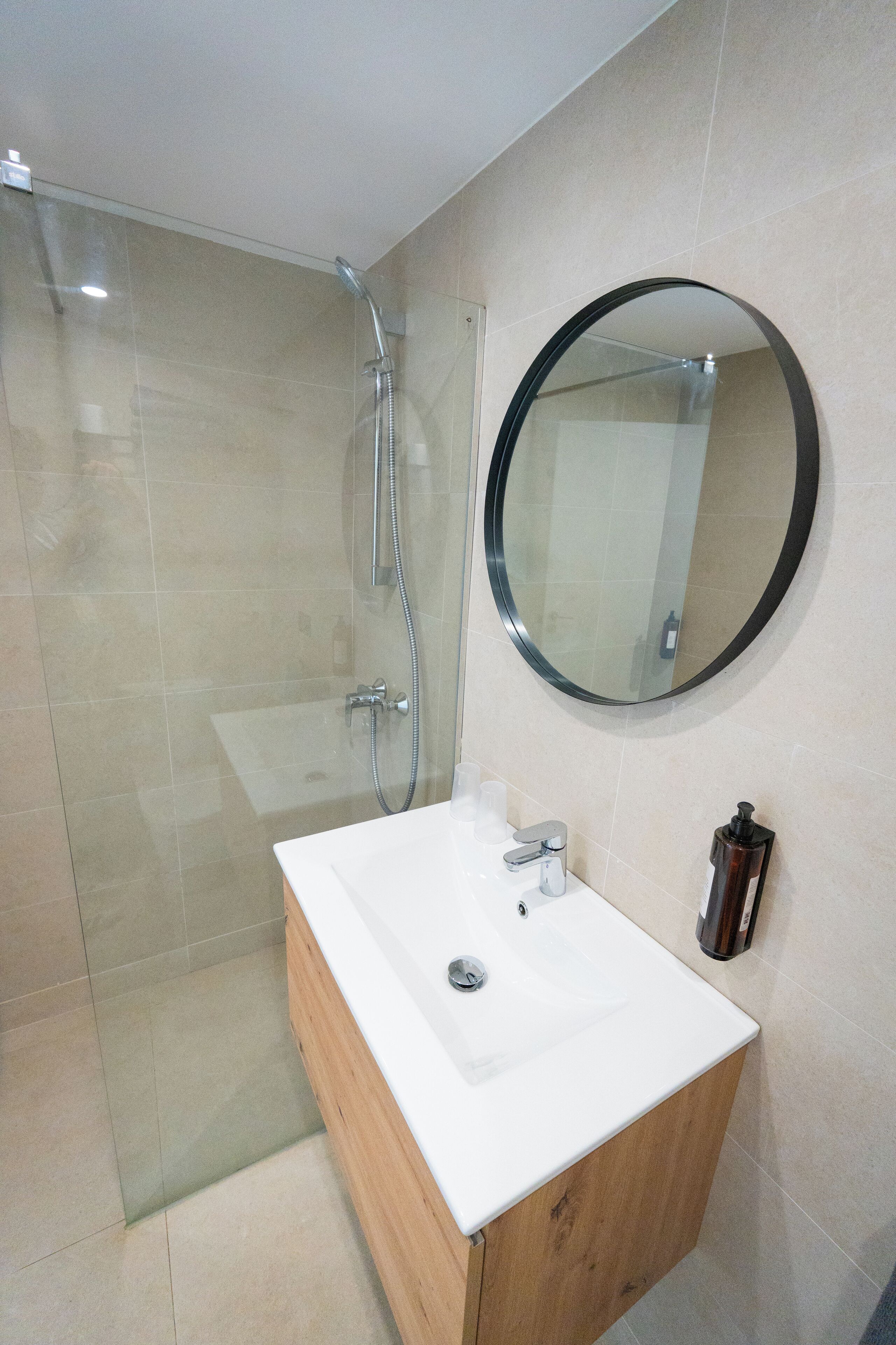 A modern bathroom with a glass shower enclosure, a white sink with a wooden cabinet, a round mirror, and a soap dispenser.