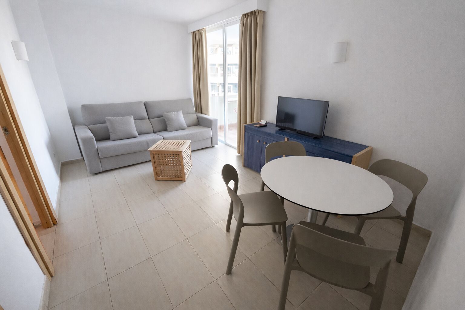 Een moderne woonkamer met een grijze bank, een houten bijzettafeltje, een ronde eettafel met vier stoelen, een televisie op een blauwe kast en grote ramen met gordijnen.