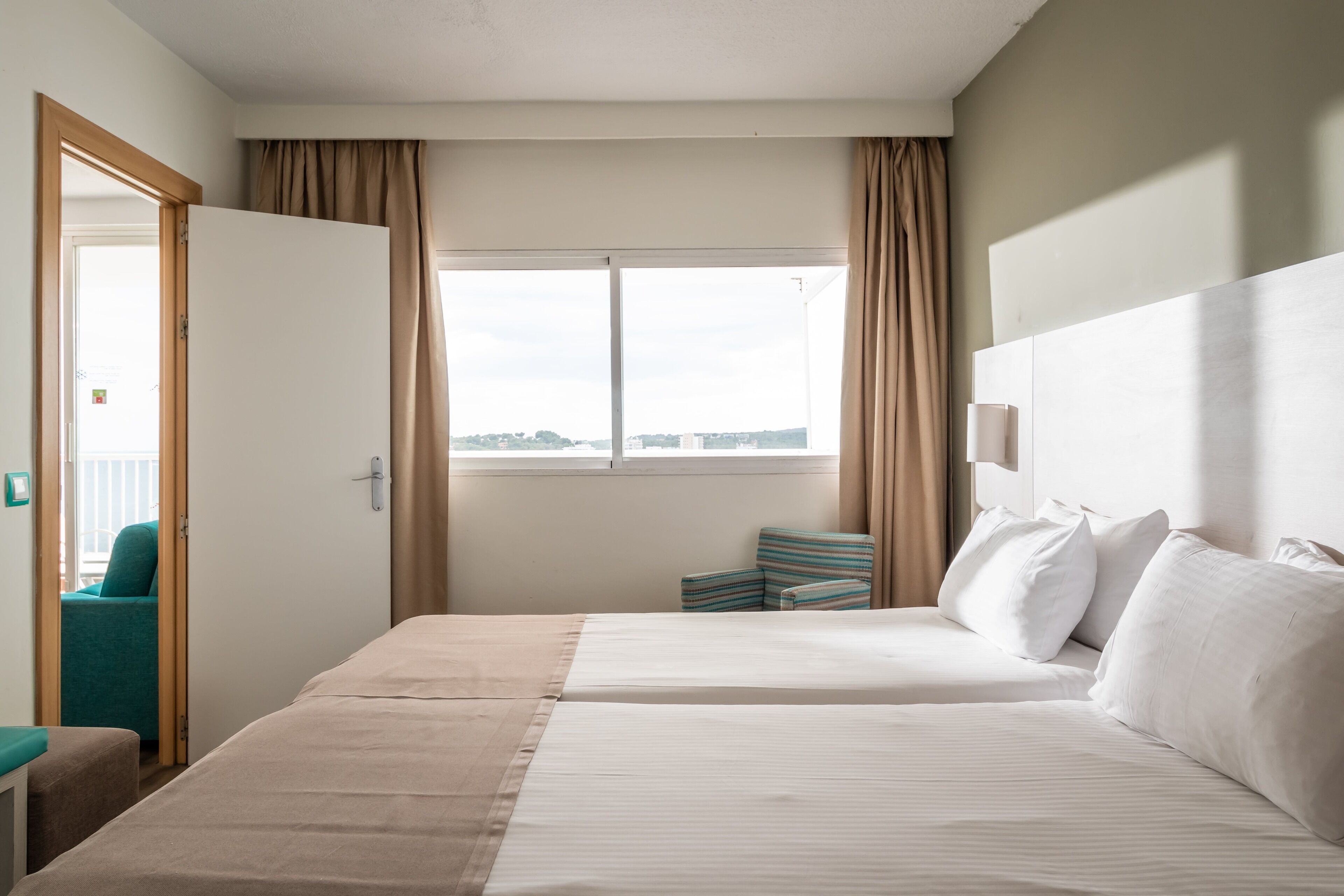 A hotel room with two beds, a view of the city, a window with curtains, and a door leading to another room.