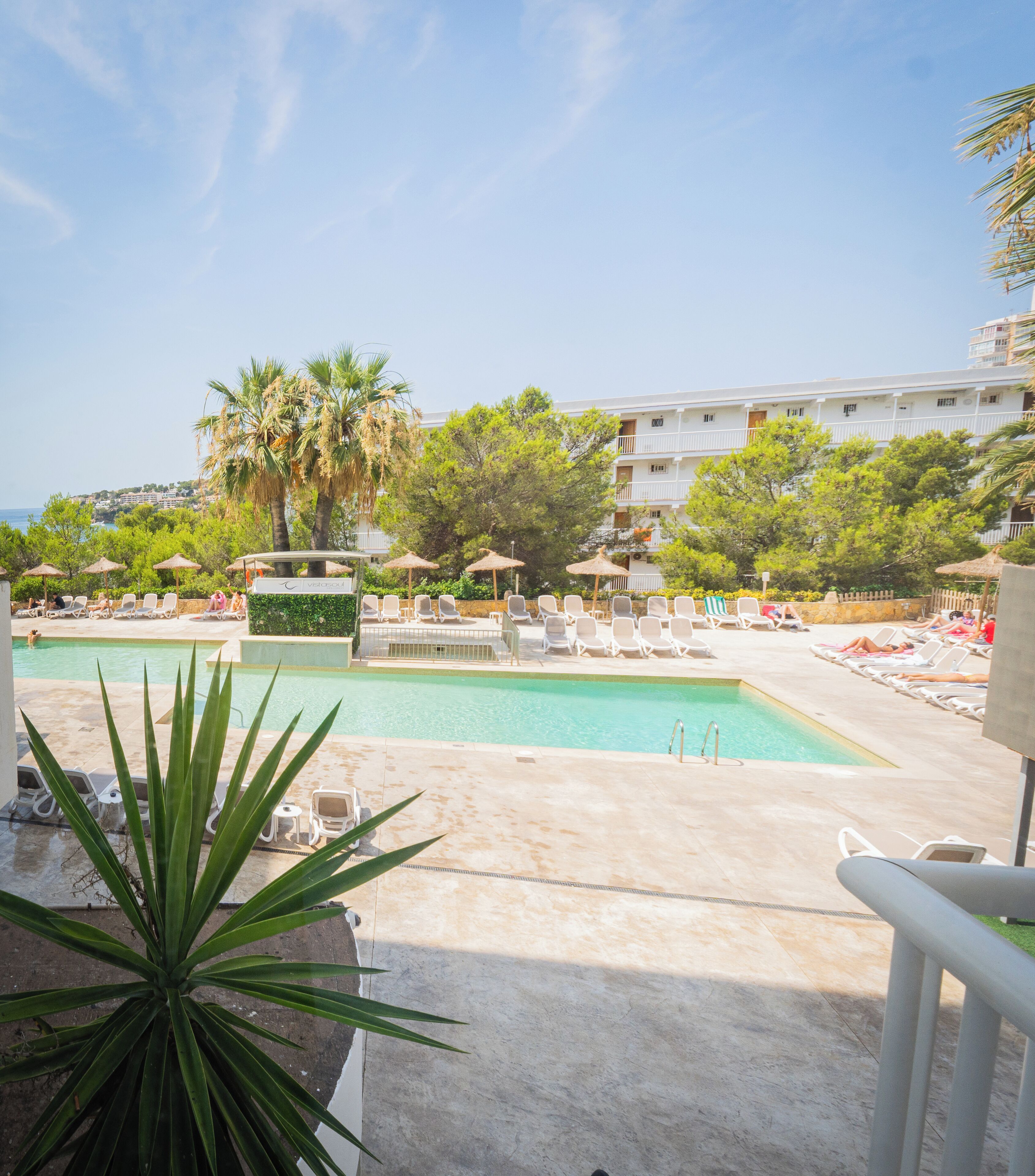 Un área de piscina con sillones de descanso y sombrillas, una palmera y un edificio al fondo.