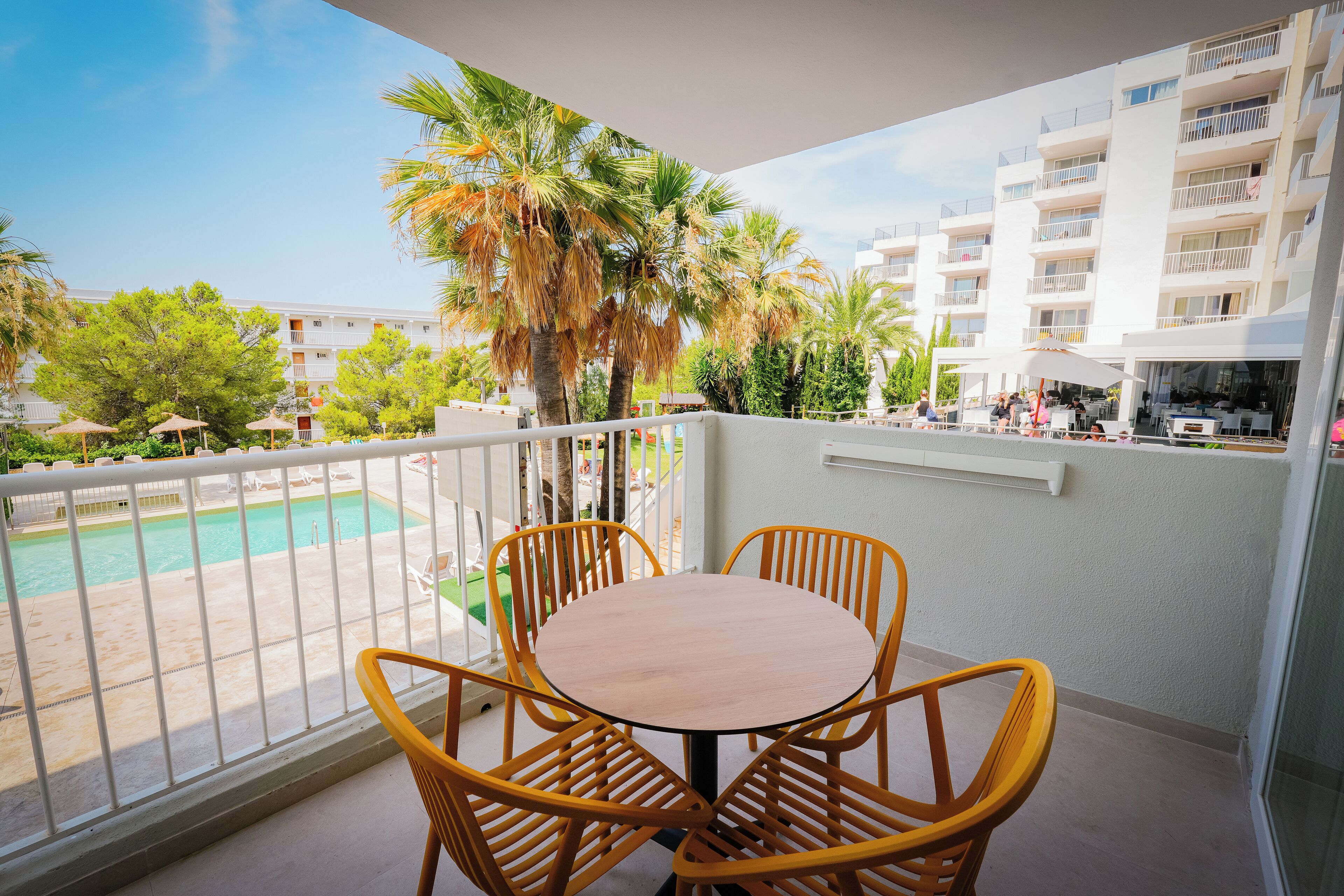 A balcony with a round table and four chairs, overlooking a swimming pool and a building with balconies.